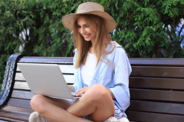 Yazlık giysiler giyen çekici genç bir kadın şehirde bankta otururken dizüstü bilgisayarla çalışıyor..