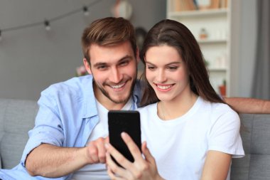 Genç çift evdeki kanepede oturmuş akıllı bir telefonla online içeriği izliyorlar..