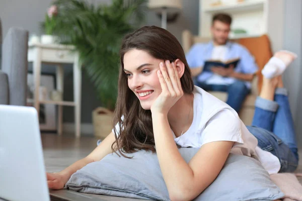 Sonriendo hermosa mujer usando el ordenador portátil con el hombre borroso en el fondo en casa . — Foto de Stock