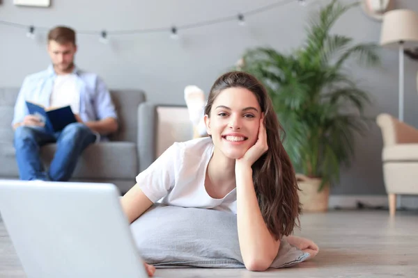 Glimlachende mooie vrouw met behulp van laptop met wazig man in de achtergrond thuis. — Stockfoto