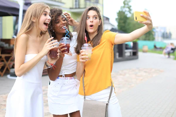 Trzy modne hipsterskie dziewczyny, przyjaciele piją koktajl i robią selfie w tle miasta. — Zdjęcie stockowe