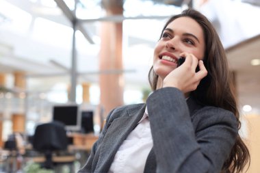 Ofiste bilgisayarın yanında akıllı telefon kullanan gülümseyen genç bir iş kadını..