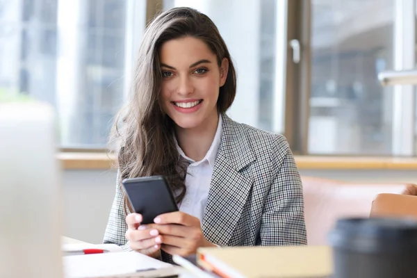 Jeune femme d'affaires souriante utilisant un smartphone près de l'ordinateur au bureau. — Photo