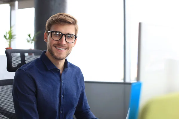 Bello sguardo giovane uomo d'affari guardando la fotocamera e sorridendo mentre seduto in ufficio . — Foto Stock