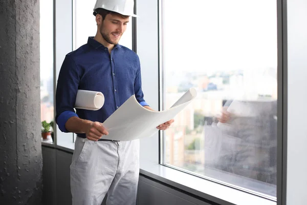 Självsäker ung affärsman i hård hatt håller ritning och tittar bort när du står inomhus. — Stockfoto