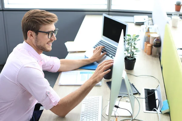 Figyelmes fiatal üzletember ingben, aki számítógéppel dolgozik, miközben az irodában áll. — Stock Fotó