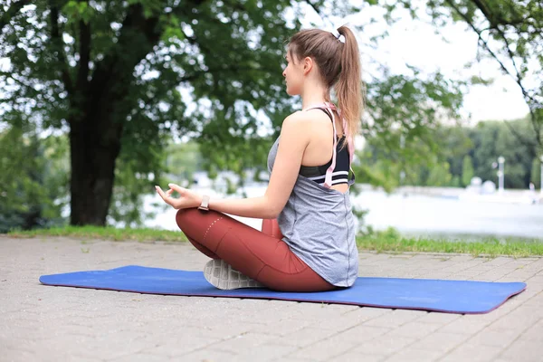 Eşofmanlı neşeli, zayıf kadın mindere oturmuş Lotus pozunda yoga yapıyor parkta sakin duruyor.. — Stok fotoğraf