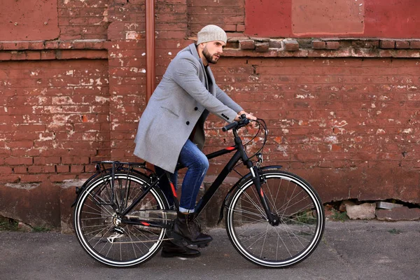 Όμορφος νέος άντρας με γκρι παλτό και καπέλο που στέκεται με το ποδήλατό του. — Φωτογραφία Αρχείου