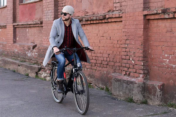 灰色のコートと帽子をかぶったハンサムな若者が市内の自転車通りに乗る. — ストック写真