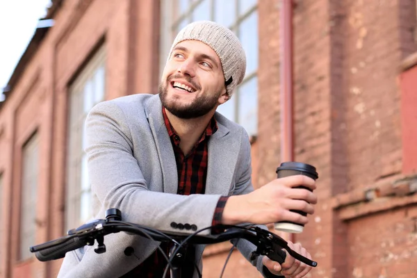 灰色のコートと帽子を着たハンサムな若者と自転車に乗ってコーヒーを飲んで通りを飲む. — ストック写真