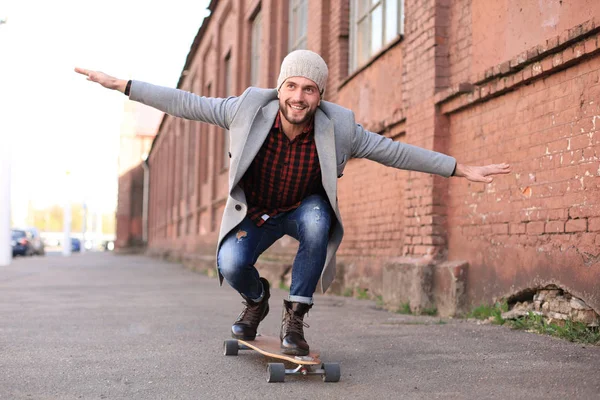 Şehirde sokakta uzun tahta üzerinde gri ceket ve şapka yakışıklı genç adam. Kentsel kaykay konsepti.