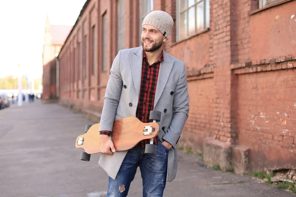 Bello giovanotto in cappotto grigio e cappello che cammina per strada, usando il longboard . — Foto Stock