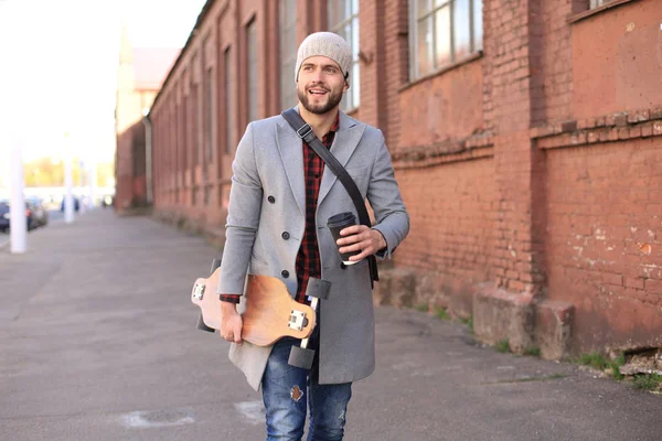 Красивий молодий чоловік у сірому пальто і капелюсі, що йде по вулиці, використовуючи дошку . — стокове фото