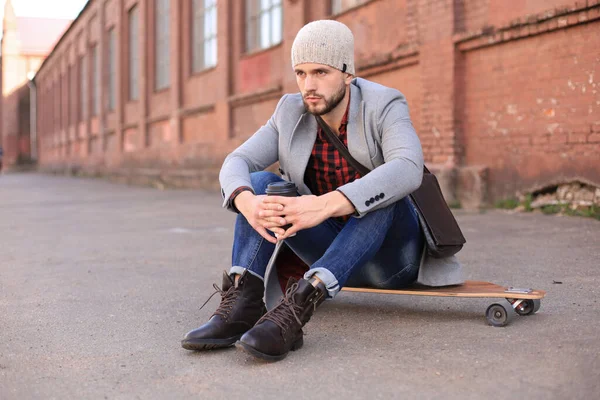 Beau jeune homme en manteau gris et chapeau assis sur le longboard dans la rue de la ville. Concept de skateboard urbain . — Photo