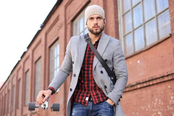 Joven guapo con abrigo gris y sombrero caminando por la calle, usando longboard . — Foto de Stock