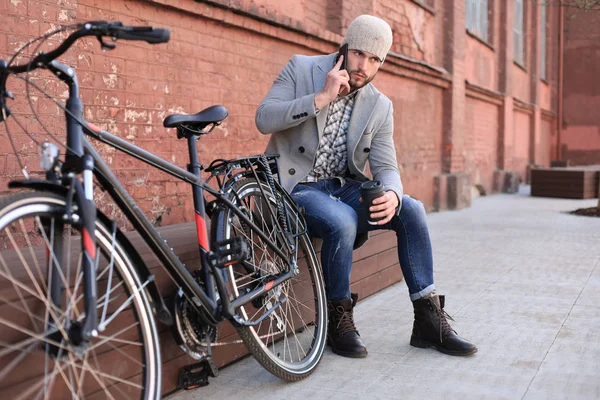 Schöner junger Mann in grauem Mantel und Hut, der mit dem Handy telefoniert und lächelt, während er im Freien neben seinem Fahrrad sitzt. — Stockfoto