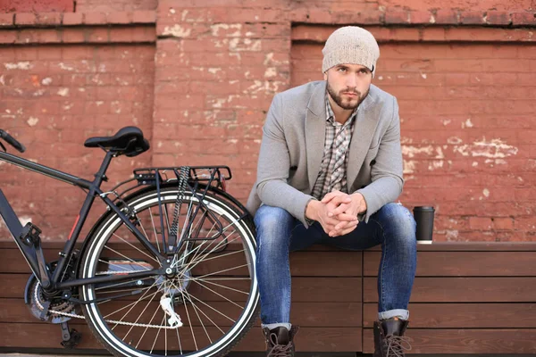灰色のコートと帽子をかぶったハンサムな若者がベンチに座ってコーヒーを飲み、自転車の近くで考えている. — ストック写真