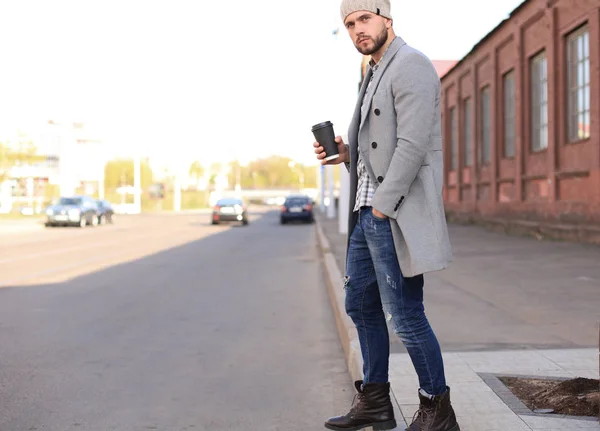 Bello giovanotto in cappotto grigio e cappello che attraversa la strada con una tazza di caffè . — Foto Stock