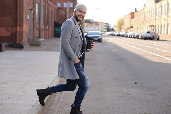 Joven guapo con abrigo gris y sombrero cruzando la calle con una taza de café . — Foto de Stock