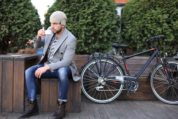 Guapo joven con abrigo gris y sombrero sentado en un banco relajado bebiendo café y pensando cerca de su bicicleta . — Foto de Stock