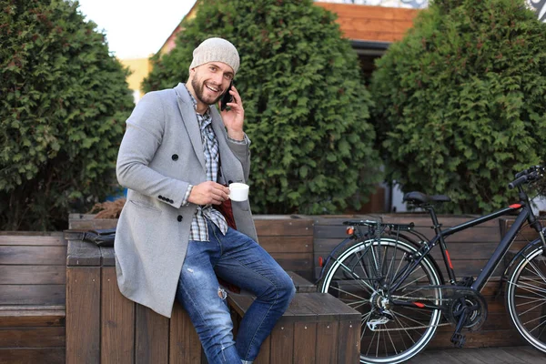 Schöner junger Mann in grauem Mantel und Hut, der mit dem Handy telefoniert und lächelt, während er im Freien neben seinem Fahrrad sitzt. — Stockfoto