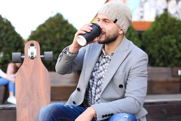 Bello giovane in cappotto grigio e cappello, a riposo, seduto con longboard bere caffè. Concetto di skateboard urbano . — Foto Stock