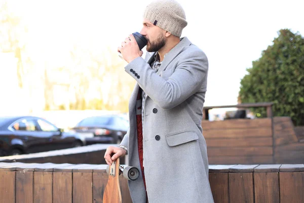 Schöner junger Mann in grauem Mantel und Hut, ruht sich aus, sitzt mit Longboard und trinkt Kaffee. städtisches Skateboarding-Konzept. — Stockfoto