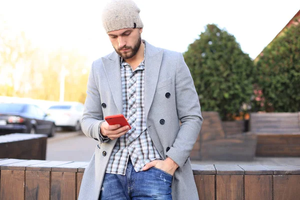 Beau jeune homme en manteau gris et chapeau avec smartphone, reposant, debout sur la rue de la ville. Concept urbain . — Photo