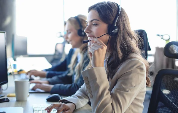 Operator dukungan pelanggan perempuan dengan headset dan tersenyum, dengan kolega di latar belakang. — Stok Foto