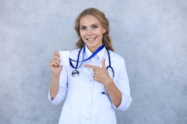 Usmívající se blondýnka lékař v uniformě stojící izolovaně nad šedým pozadím, ukazující své jméno na odznaku. — Stock fotografie