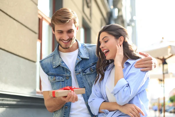 Astonished excited couple in summer clothes smiling and holding present box together while standing on city street.