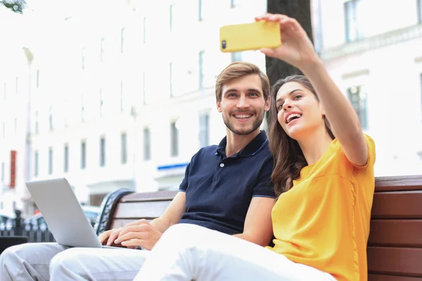 Atractiva pareja joven sentada en un banco y tomando selfie en el teléfono inteligente . — Foto de Stock