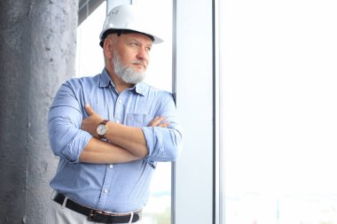 Thoughtful mature architect wearing hardhat and looking away while standing near the window in new modern building.