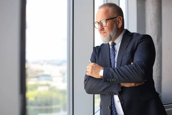 Dospělý obchodník v podnikovém obleku, který stojí v úřadu a velmi optimisticky hledí do velkých oken. — Stock fotografie