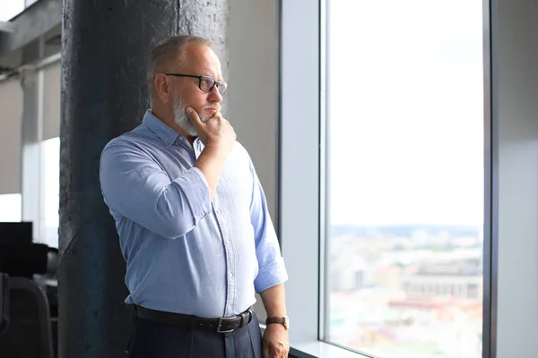 Gondolkodó érett üzletember egy vállalati öltönyt néz el, miközben áll az ablak közelében. — Stock Fotó
