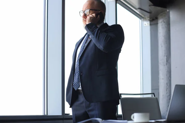 Зрілий бізнесмен розмовляє на мобільному телефоні, стоячи біля вікна з видом на місто . — стокове фото