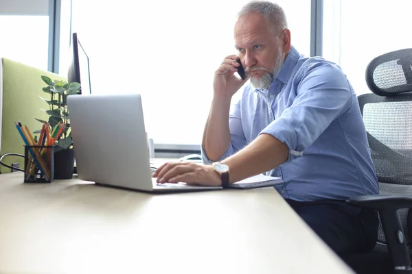 Selbstbewusster, reifer Geschäftsmann, der im modernen Büro mit seinem Smartphone telefoniert. — Stockfoto