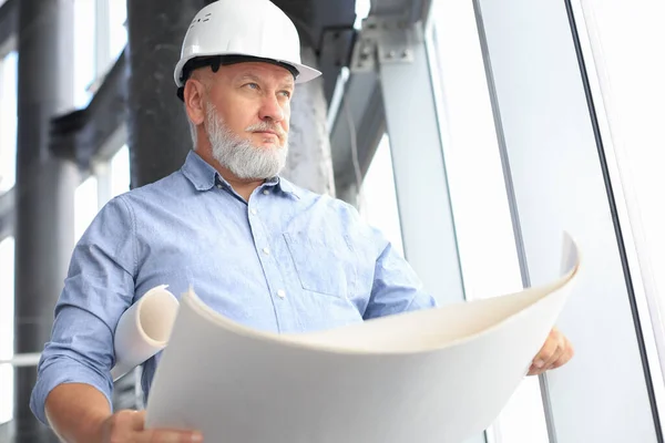 Confident mature architect in corporate suit and hardhat holding a blueprint and looking at it.