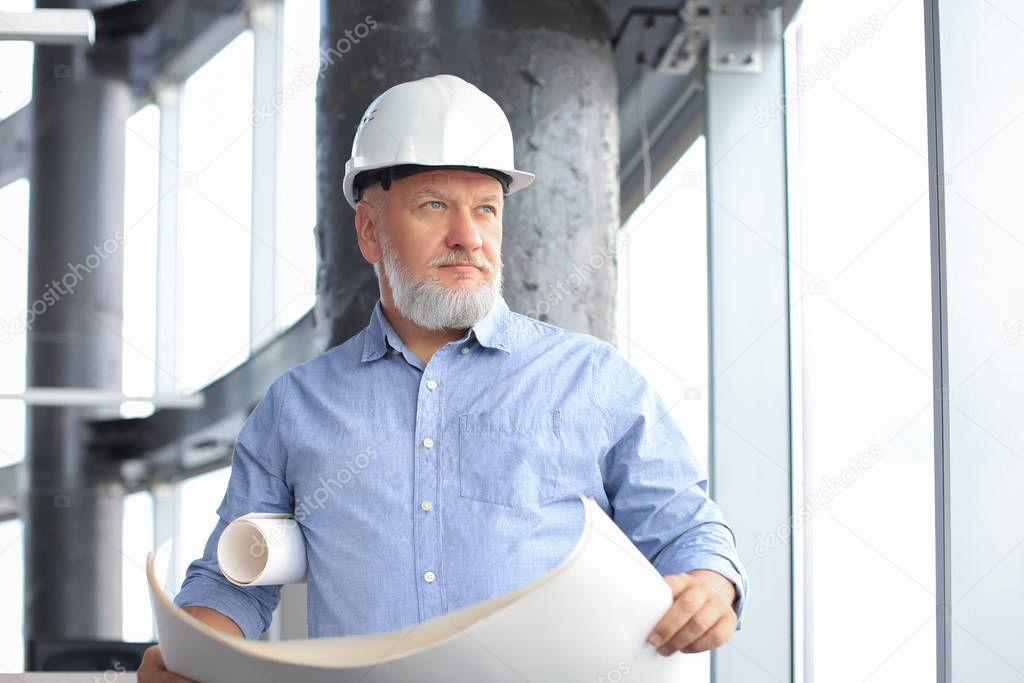 Confident mature architect in corporate suit and hardhat holding a blueprint and looking at it.