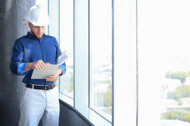 Kasketli, kendine güveni tam genç bir iş adamı elinde planlar ve içeride duran belgelere bakıyor..