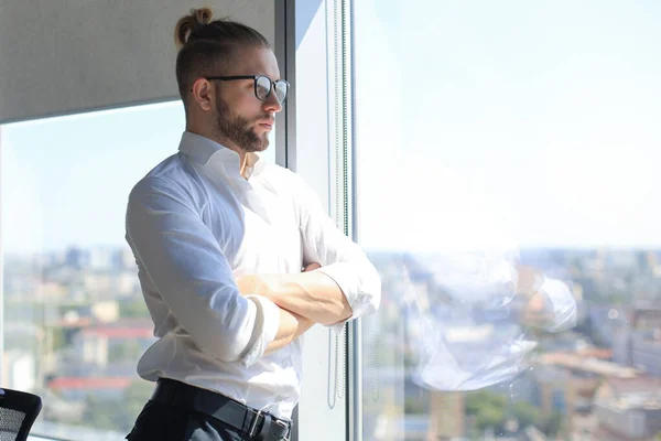 Joven hombre de negocios serio manteniendo los brazos cruzados y mirando hacia otro lado mientras está de pie cerca de la ventana . —  Fotos de Stock