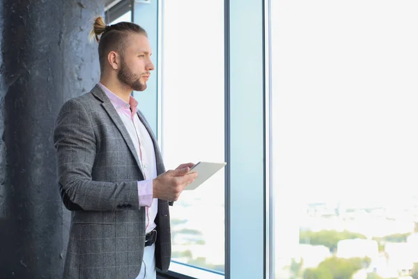 Guapo joven empresario sosteniendo tableta digital mientras está de pie cerca de la ventana . — Foto de Stock