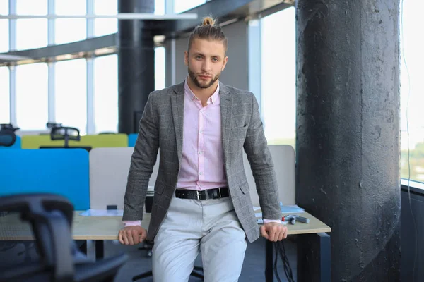 Продуманий молодий бізнесмен в повсякденному одязі дивиться на камеру і тримає руку на підборідді, спираючись на стіл . — стокове фото