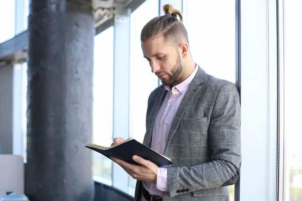 Goed uitziende jonge zakenman in casual kleding die zijn notitieblok leest terwijl hij in het kantoor staat. — Stockfoto