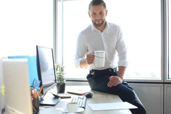 Dobře vypadající mladý podnikatel pije kávu a usmívá se, zatímco sedí v kanceláři. — Stock fotografie