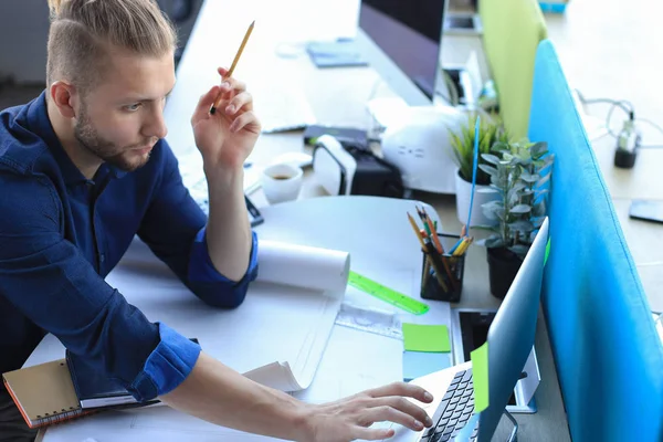 Fotografía de un atractivo arquitecto masculino sosteniendo planos mientras está sentado en su oficina . — Foto de Stock