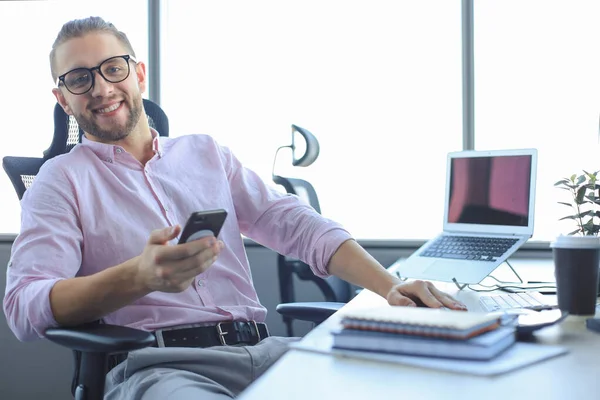 スマートフォンを使いオフィスに座っている間に笑顔で若いビジネスマンをよく見る. — ストック写真