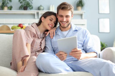 Pijamalı genç bir çift, oturma odasındaki kanepede oturmuş tablette medyanın içeriğini izliyorlar..