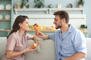 Mutfakta pijamalarıyla birbirlerini kruvasanla besleyen mutlu genç bir çift..