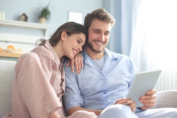 Pareja joven en pijama viendo contenido multimedia en línea en una tableta sentada en un sofá en la sala de estar. —  Fotos de Stock